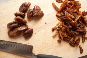 Wooden chopping board with a pile of chopped dates on one side, and a pile of unchopped dates on the other. Below the dates is a sharp knife.
