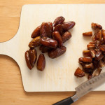 Chopping board with unchopped dates on one half and chopped dates on the other half. A knife is near the bottom.