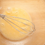 Top-down view of beaten egg in a glass bowl with a balloon whisk resting in the bowl. The bowl is on a wooden table.