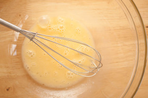 Top-down view of beaten egg in a glass bowl with a balloon whisk resting in the bowl. The bowl is on a wooden table.